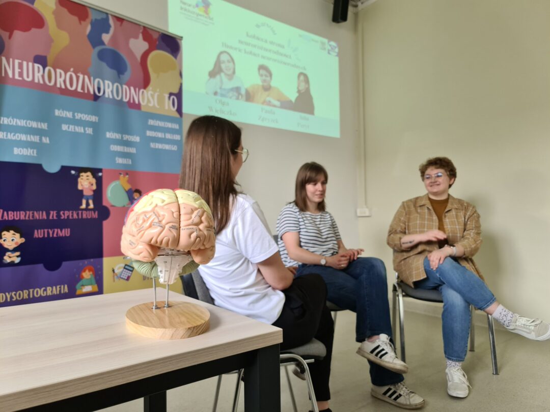 Zdjęcie przedstawia trzy dziewczyny, siedzą na krzesłach. Opowiadają one o swoich doświadczeniach. Za nimi jest prezentacja multimedialna.
